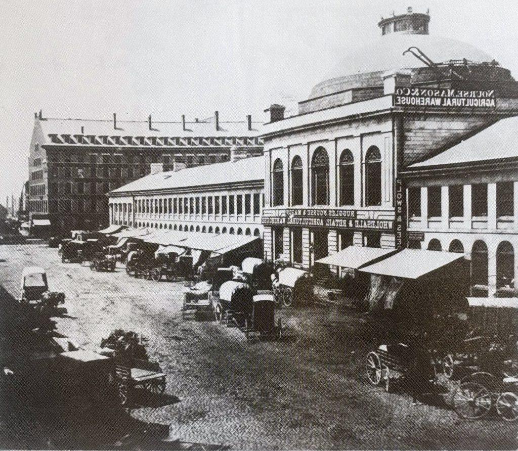 Next to the large stone side of Quincy Market, a street with market tents and covered wagons.