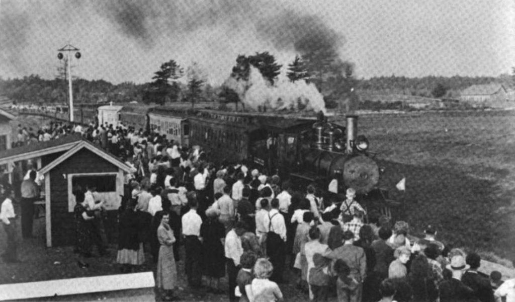 Black and white photograph of a crowd of people standing outside next to a train pulling into a station. The train engine pulls several cars, and smoke billows out of its chimney. Behind the train and at the left of the picture are fields, trees, and farm buildings. At the right is a small station building and a railroad signal on a pole.